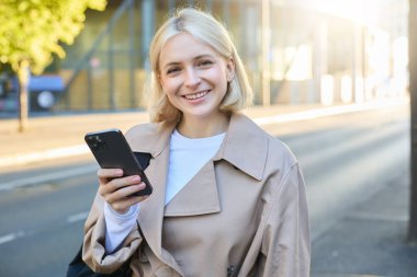 Gülümseyen sarışın kadın portresi, sokakta trençkot giyiyor, mutlu görünüyor, cep telefonu kullanıyor, akıllı telefon kullanıyor, memnun ve tatmin olmuş bir yüz ifadesi var..