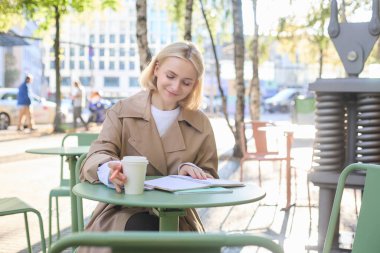 Genç gülümseyen kadın portresi, açık havada çalışmak, kafede defter ile oturmak, yazmak, kahve içmek, dinlendirici atmosferde temiz hava üzerine ödev yapan öğrenci..