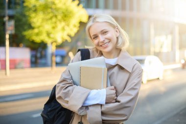 Sarışın, gülümseyen güzel bir kadın resmi güneşli bir günde caddede yürüyor, defterlerini ve üniversite malzemelerini taşıyor, tasasız görünüyor..