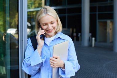 Sırtında dizüstü bilgisayarı olan, sırtüstü akıllı telefondan konuşan, bir telefona cevap veren, gülümseyen sarışın kadının yakın portresi..