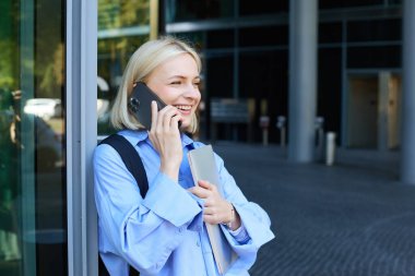 Mutlu kadın model, sarışın kadın cep telefonuyla konuşuyor, biriyle sohbet ediyor, ofis binasının yanında dizüstü bilgisayarı ve akıllı telefonuyla duruyor, gülümsüyor..