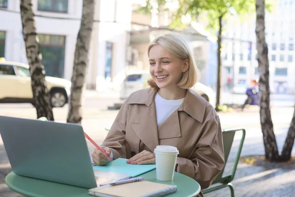 Remote workplace. Smiling young woman freelancer, student using laptop in outdoor cafe, talking to someone, attends online lecture, drinking coffee.