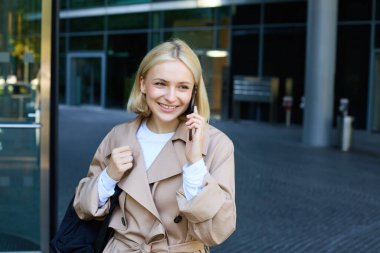 Akıllı telefonlu kaygısız genç bir kadın, şehir merkezinde dışarıda yürüyor, caddede duruyor ve sohbet ediyor, cep telefonuyla konuşuyor, mutlu, güler yüzlü bir yüz ifadesiyle..