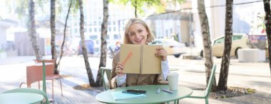 Tasasız, gülümseyen genç bir kadının portresi defter ve kalemle, açık kafede masa başında oturuyor, günlüğe yazıyor, ajandasında planlar yapıyor..
