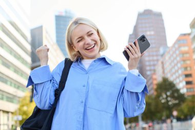 Sırt çantası ve cep telefonu taşıyan mutlu, hevesli genç bir kadının portresi, caddede durur ve sevinir, zafer kazanır, yumruk hareketi yapar, mutluluk ve neşe içinde dans eder..