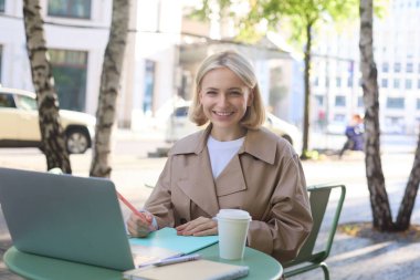 Sokakta oturan, kafede kahve içen, bilgisayarla uzaktan çalışan, online kursa katılan ya da ev ödevi yapan genç bir kadının resmi..
