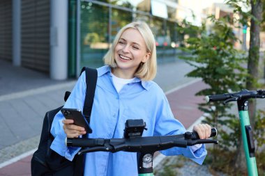 Gülümseyen sarışın kadın portresi, akıllı telefon uygulaması kullanan üniversite öğrencisi yeşil elektrikli scooter 'da QR kodunu tarıyor, araba kiralıyor..