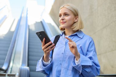 Mavi gömlekli güzel sarışın bir kadının portresi yürüyen merdivenin yanında duruyor, cep telefonunu tutuyor, akıllı telefon kullanıyor, şehir merkezinde birini bekliyor..