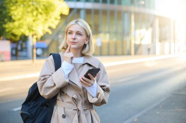 Düşünen yüzlü bir kadın portresi, sırt çantasıyla sokakta duruyor, akıllı telefon tutuyor, düşünüyor, düşünceli bir yüzle karar veriyor..