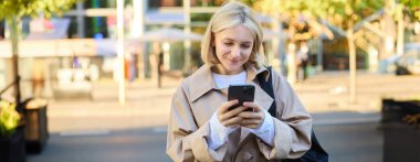 Sokaktaki modern genç kadın, cep telefonundan mesaj okuyor, akıllı telefon kullanıyor, yoğun yolun yanında duruyor ve gülümsüyor..