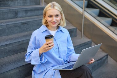 Sarışın, gülümseyen bir kadının portresi, kahve içen, dizüstü bilgisayar kullanan, binanın dışındaki kampüs merdivenlerinde oturan, okuyan, e-öğrenen, kablosuz internete bağlanan..