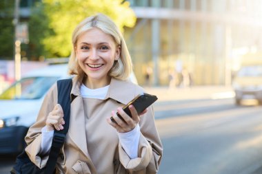 Genç modern kadının portresi, sokakta dikilen, cep telefonlu üniversite öğrencisi, kameraya mutlu ve gülümsüyor, akıllı telefon uygulaması kullanıyor..