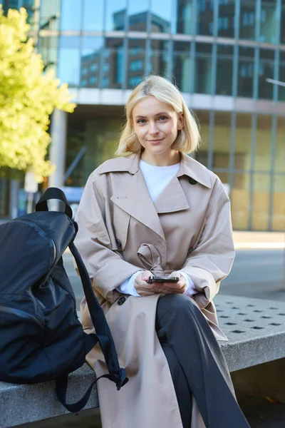 Vertical shot of young female employee, woman sitting on street bench with backpack and smartphone, waiting for her taxi, using mobile app, looking confident at camera.