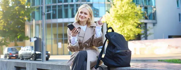 Portrait of smiling female employee, woman sitting on bench with mobile phone and backpack, pointing finger at copy space, showing promo banner, advertising.
