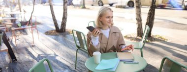 Çalışan genç bir kadının portresi, akıllı telefonlar ve günlüklerle açık bir kafede oturan, ev ödevi yapan, güneşli bir günde yağmurluk giyen.