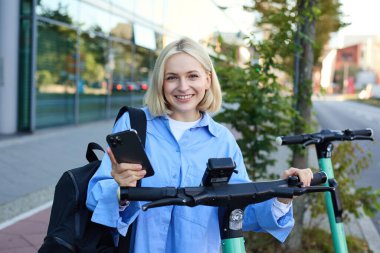 Genç gülümseyen bir kadının portresi, elektrikli scooter 'ın kilidini açıyor, akıllı telefon uygulaması ile QR kodunu tarayıp aracının kilidini açıyor, binanın yanındaki sokakta duruyor..