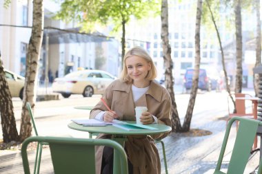 Gülümseyen, neşeli bir kadın kahvesini sokak kafesinde içiyor, deftere yazıyor, ödev yapıyor, notlar alıyor. Öğrenci ve yaşam tarzı konsepti.