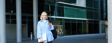 Üniversite ve insanlar konsepti. Sokakta ofis binasının yanında duran genç bir kadının dikey portresi. Üniversite kampüsünde elinde akıllı telefon, dizüstü bilgisayar ve sırt çantası olan bir öğrenci..