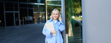 Bilgisayarlı genç bir kadının hayat tarzı portresi, kampüsün yakınında poz veriyor, ofis binasına giriyor, sırt çantası tutuyor, gülümsüyor ve mutlu görünüyor..