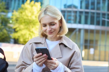 Sokakta gülümseyen kadın modelin yakın portresi, cep telefonuyla bankta oturmak, akıllı telefon ekranına bakmak, dışarıda beklerken arkadaşınla mesajlaşmak..