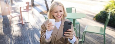 Neşeli, güzel bir genç kadının portresi selfie çekiyor, akıllı telefon kullanarak görüntülü sohbet ediyor, açık havada kahve dükkanında poz veriyor..