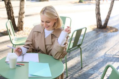 Genç konsantre bir kadının portresi, kafede ödev yapmak, deftere yazmak, belgelerle çalışmak ve dışarıda kahve içmek..