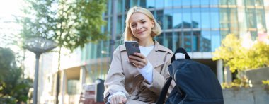 Gülümseyen genç bir kadının portresi, sokak bankında oturuyor, cep telefonunu tutuyor, dışarıda arkadaşını beklerken akıllı telefondan biriyle sohbet ediyor..