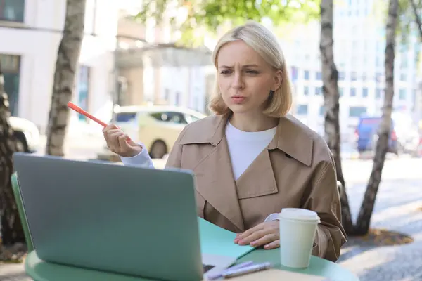 Image of woman with confused face, looking at laptop, working remotely, chatting with someone and shrugging, talking with puzzled face, sitting in coffee shop.