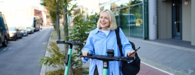 Gülümseyen sarışın kadın portresi, akıllı telefon uygulaması kullanan üniversite öğrencisi yeşil elektrikli scooter 'da QR kodunu tarıyor, araba kiralıyor..