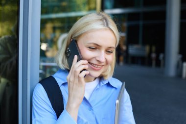 Mutlu kadın model, sarışın kadın cep telefonuyla konuşuyor, biriyle sohbet ediyor, ofis binasının yanında dizüstü bilgisayarı ve akıllı telefonuyla duruyor, gülümsüyor..