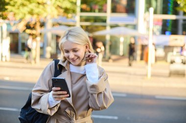 Sokaktaki modern genç kadın, cep telefonundan mesaj okuyor, akıllı telefon kullanıyor, yoğun yolun yanında duruyor ve gülümsüyor..