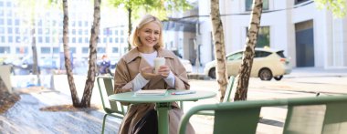 Şehir yaşam tarzı konsepti. Genç ve güzel bir kadın açık hava kafede oturuyor, şehir hayatını düşünüyor, kahve içiyor ve sokakta gülümsüyor..