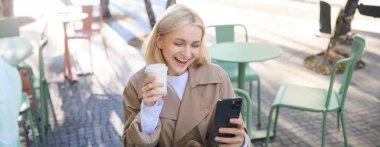 Gülümseyen genç bir kadının hayat tarzı portresi, açık havada kahve içen, mutlu, heyecanlı bir yüz ifadesiyle akıllı telefona bakan. Teknoloji ve insanlar kavramı