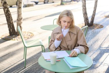 Ödev yaparken düşünen genç bir kadının portresi, açık kafede tek başına oturmak, kahve içmek ve deftere yazmak, düşünceli görünmek, üzerine kafa yormak..