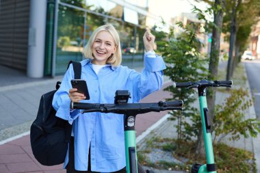 Genç, gülümseyen kadın modelin portresi, elektrikli scooter kiralamak, akıllı telefon uygulaması ile QR kodunu taramak için cep telefonu kullanmak, üniversiteden eve gitmek, sokakta dikilmek..