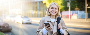 Sırt çantalı, sokakta durup cep telefonu kullanan, yol kenarında taksi bekleyen, gülümseyen mutlu ve güzel genç bir kadının portresini yakından çek..