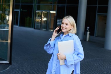 Ofiste çalışan genç bir kadın resmi, şirket binasının yanında elinde belgeler ve defterlerle duran, telefonlara cevap veren, akıllı telefondan konuşan, gülümseyen, mavi yakalı gömlekli..