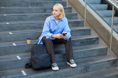 Modern bir kadının portresi, sırt çantalı bir öğrenci öğle yemeği molasında merdivenlerde oturuyor, akıllı telefon kullanıyor, yan bakıyor..