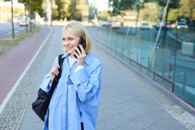 Neşeli, genç, modern bir kadın sokakta yürüyor ve telefona cevap veriyor, cep telefonuyla konuşuyor, siyah sırt çantası taşıyor..