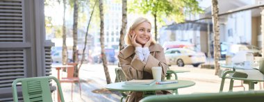 Şehir yaşam tarzı konsepti. Genç ve güzel bir kadın açık hava kafede oturuyor, şehir hayatını düşünüyor, kahve içiyor ve sokakta gülümsüyor..