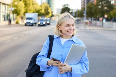 Mavi tişörtlü, sırt çantalı ve akıllı telefonlu sarışın bir kadının portresi. Arabasını bekliyor, taksi çağırıyor, cep telefonundan güncellemeleri kontrol ediyor, caddenin yanında duruyor..