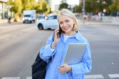 Mavi gömlekli modern kadın modelin portresi, cep telefonuyla konuşmak, sokakta yürürken biriyle sohbet etmek, dizüstü bilgisayar tutmak. Yaşam tarzı ve insan kavramı