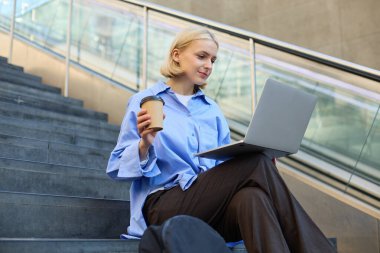 Modern, şık bir genç kadın, ev ödevi yapan bir öğrenci, kampüs merdivenlerinde çalışıyor, laptopla ve kahveyle oturuyor, cappuccinosunu içiyor ve halka açık kablosuz internet bağlantısı kuruyor..