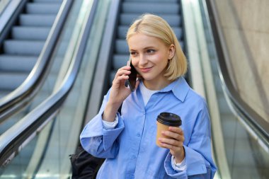 Yakışıklı sarışın bir kadının portresi, kahve fincanı olan bir öğrenci, telefona cevap veriyor, şehirde yürüyen merdivenin yanında duruyor, akıllı telefondan biriyle konuşuyor, sohbet ediyor..