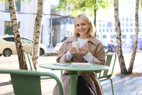 Концепция городского образа жизни. Молодая красивая женщина сидит в кафе на открытом воздухе, созерцает городскую жизнь, пьет кофе и улыбается на улице.