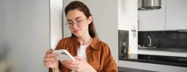 Evdeki genç kadının portresi, akıllı telefondan haberleri veya mesajları kontrol etmek, online alışveriş, cep telefonu uygulamasından teslimatı onaylamak, duvara yaslanmak, oturma odasında durmak..