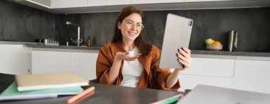 Evden çalışan, mutfakta oturan ve ekiple konuşan genç bir kadının portresi çevrimiçi toplantı yapıyor. Webinar 'a katılmak için dijital tablet kullanıyor, sohbet ediyor..
