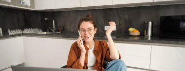 Dizüstü bilgisayarın önünde cep telefonuyla konuşan, mutlu bir yüz ifadesiyle mutfakta oturan, sohbet eden heyecanlı genç bir kadın..