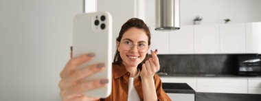 Güzel genç bir kadın, sosyal medya blogcusu video kaydı, online video sohbeti akıllı telefondan, cep telefonuyla evde duruyor..