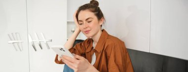 Genç bir kadın mutfak tezgahında oturuyor ve akıllı telefonunu kullanıyor. Sosyal medya, boş zaman ve mobil bağlantı kavramı.